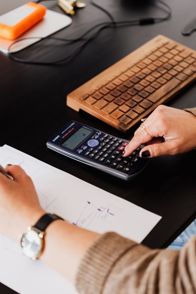 Person using calculator and writing notes on paper near wooden keyboard at desk for pool design engineering