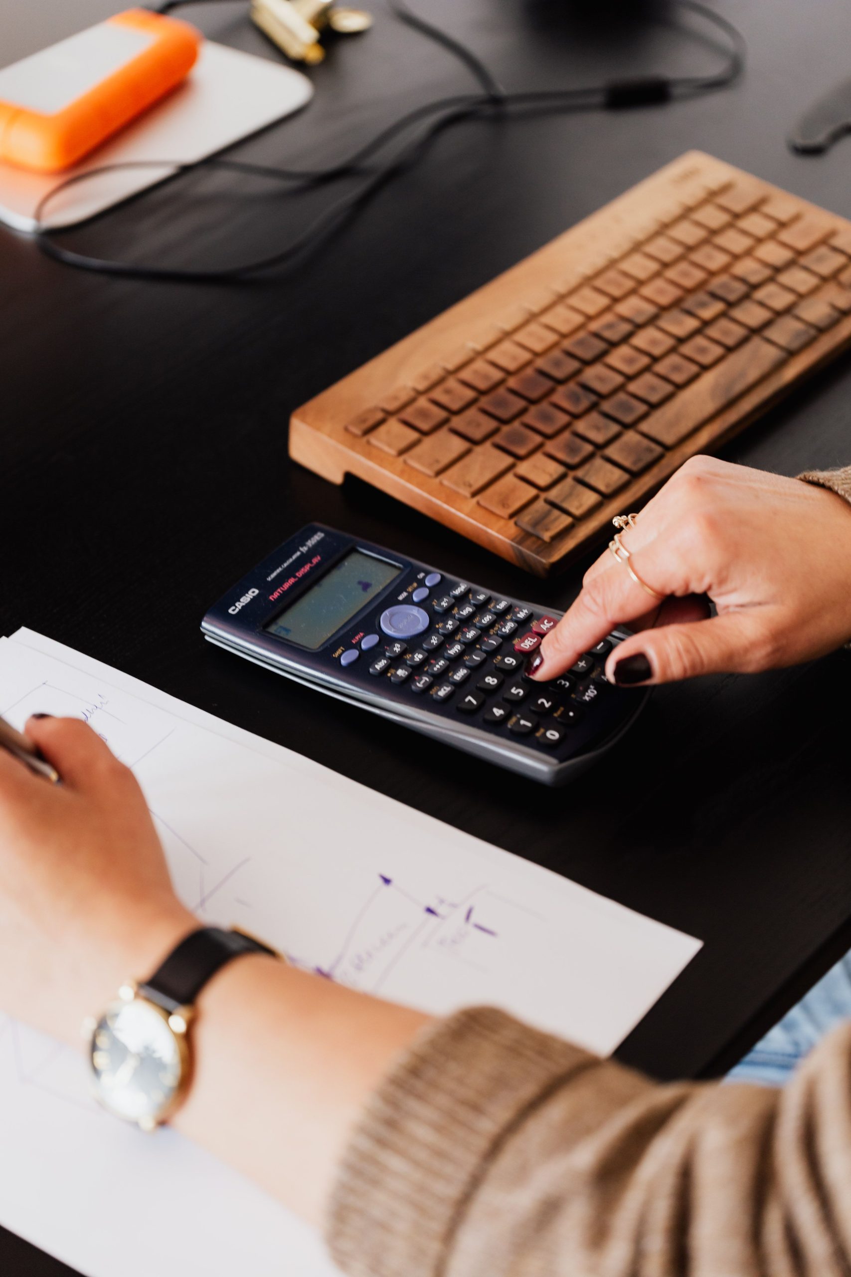 Person using calculator and writing notes on paper near wooden keyboard at desk for pool design engineering